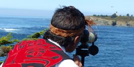A Makah Elder with red and black regalia looks through a spotting scope towards Tatoosh Island and the lighthouse from Cape Flattery