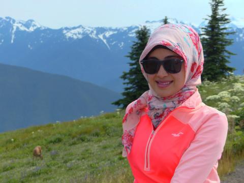 A woman with sunglasses smiles with snowclad mountains and a black-tailed deer browsing in a subalpine meadow